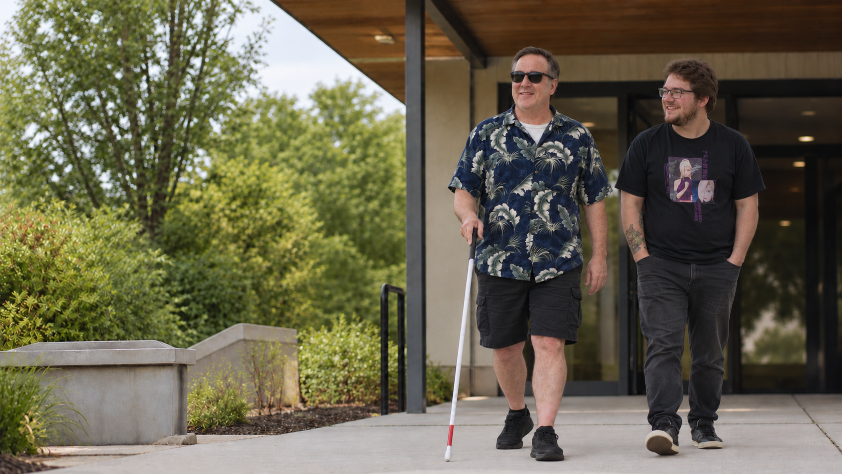Two men walking outdoors, one using a white cane.